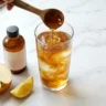 Apple cider vinegar being poured into a glass of water with raw ACV bottle and apple on marble surface