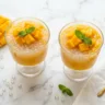 Two glasses of creamy mango sago dessert with tapioca pearls and fresh mango chunks on white marble surface