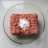 Raw ground beef in a bowl with baking soda sprinkled on top and a measuring spoon nearby