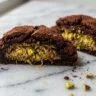 A chewy chocolate cookie cut in half showing golden pistachio kataifi filling