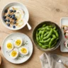 An assortment of high protein snacks including yogurt, eggs, and edamame arranged on a table