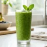 A vibrant green smoothie in a tall glass with condensation, garnished with mint