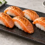 Lightly seared salmon nigiri on a dark rectangular plate with soy sauce