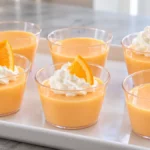 Pale orange creamsicle jello shots in small cups with optional whipped cream and orange garnish